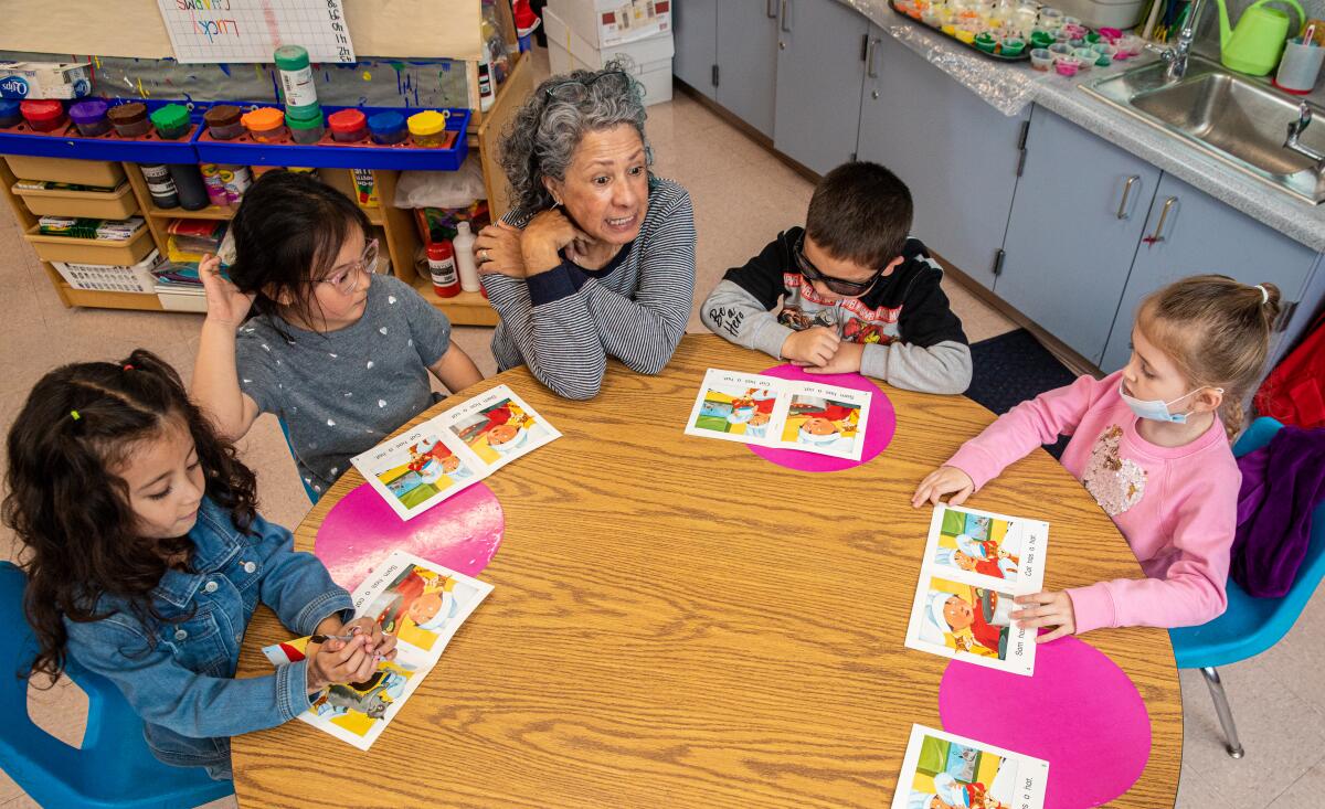 A teacher sits at a round table surrounded by four transitional kindergarteners as they work on reading.