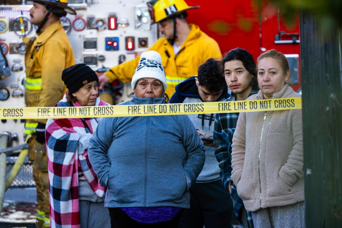 A small group standing behind a yellow ribbon reading "The line of fire does not cross" as a crew works from a fire truck in the background