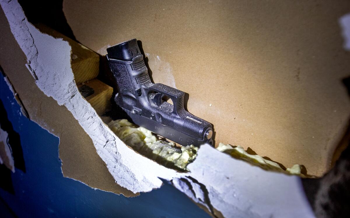 A handgun sits in a cavity behind broken drywall.