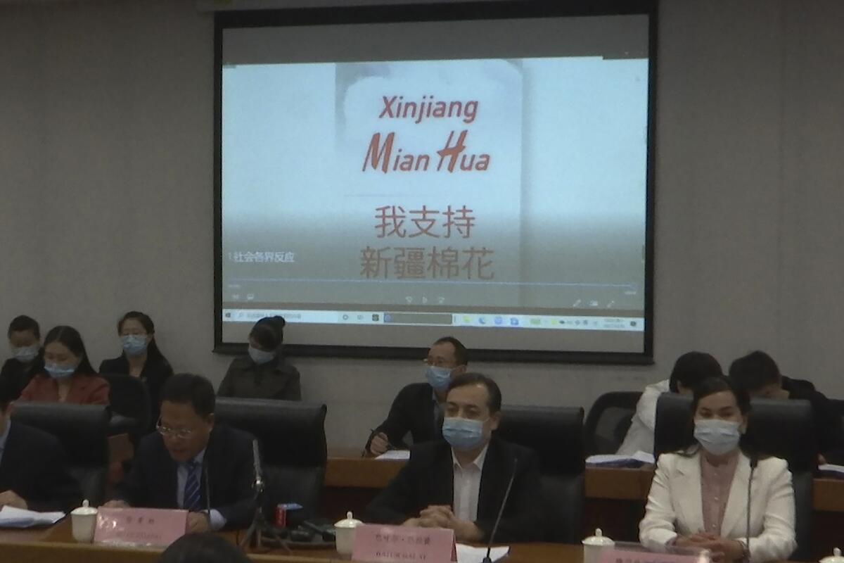 Masked people seated in front of a screen bearing the words 'Xinjiang Mian Hua.'