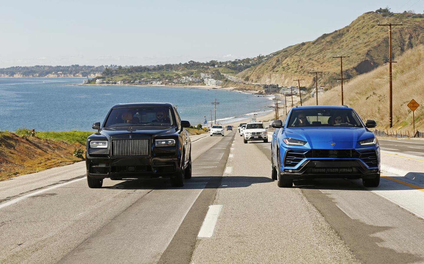 The Lamborghini Urus, in blue, and the Rolls-Royce Cullinan, in black, side by side on a Malibu road.