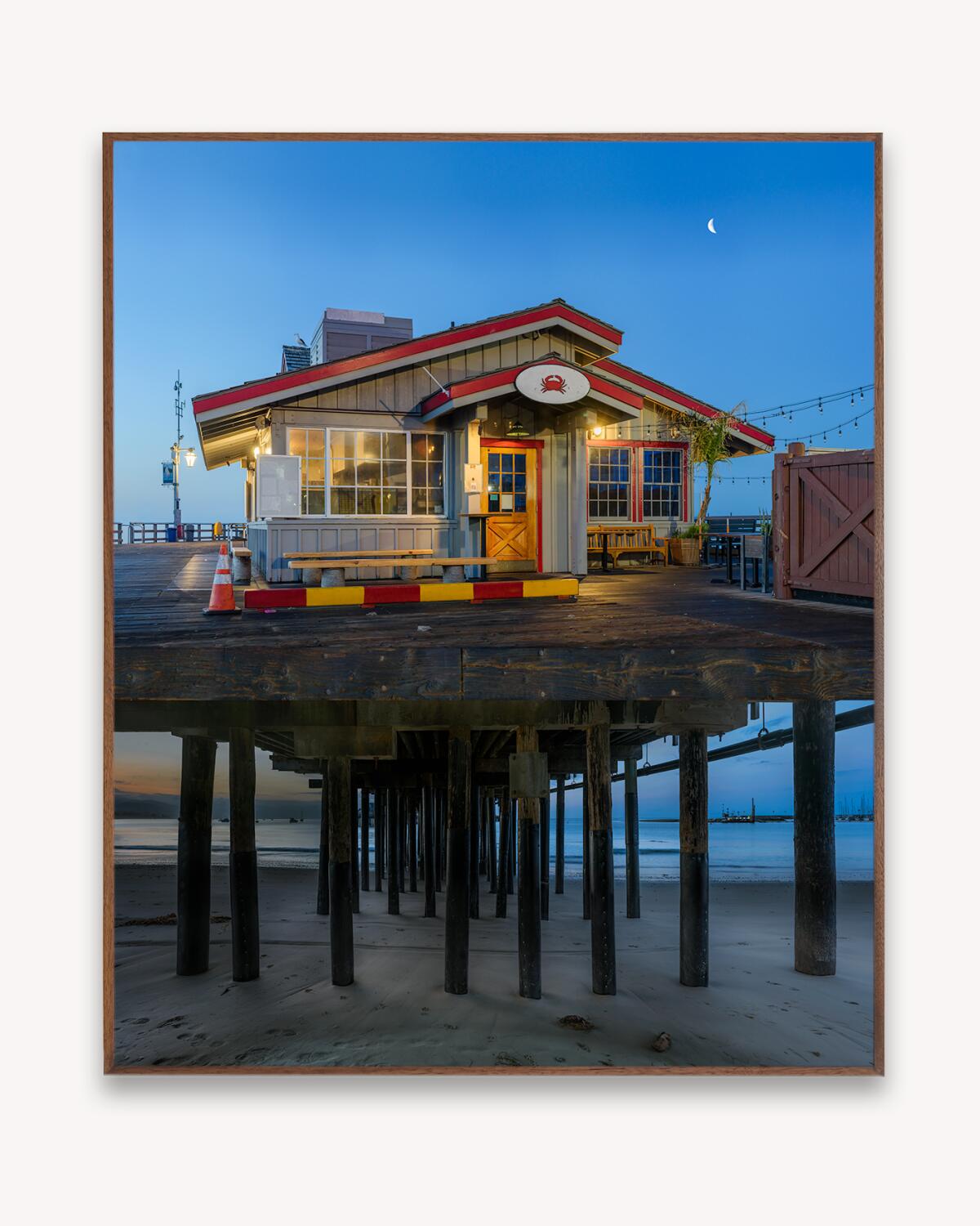 A photo of a pier and its pilings. Atop is a small building with a red roof.