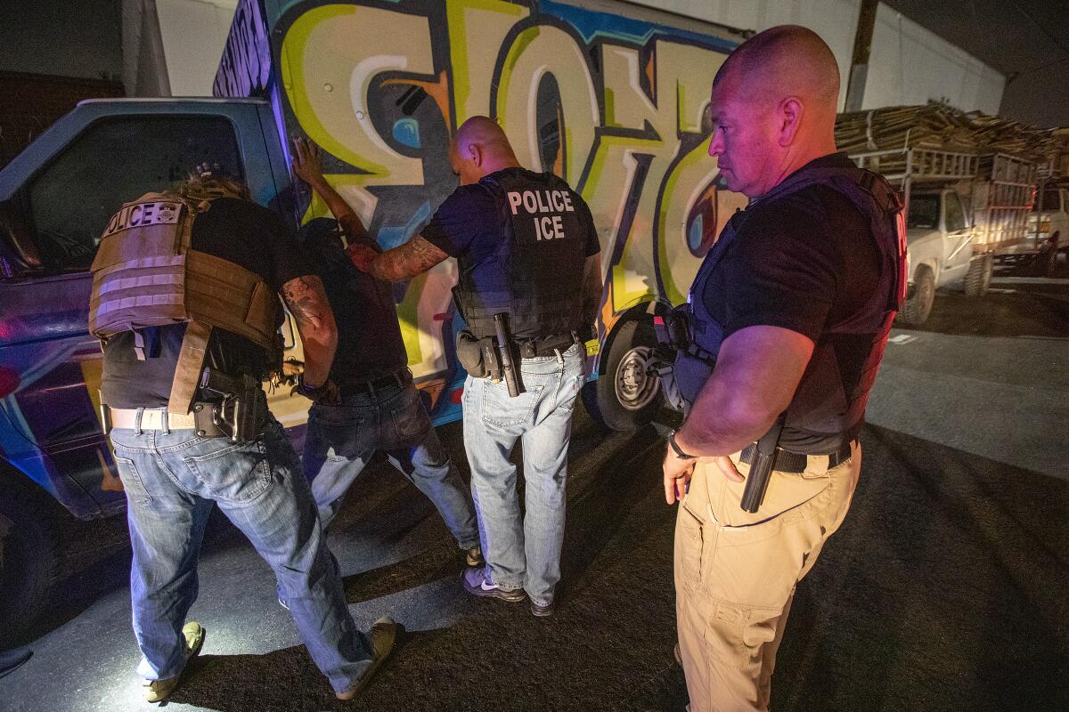 Two men put their hands on a man with his hands on a wall as another man watches.