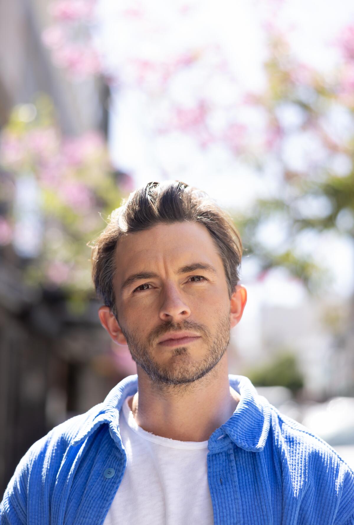 The actor Jonathan Bailey in a blue shirt over a white shirt, standing before a flowering tree.