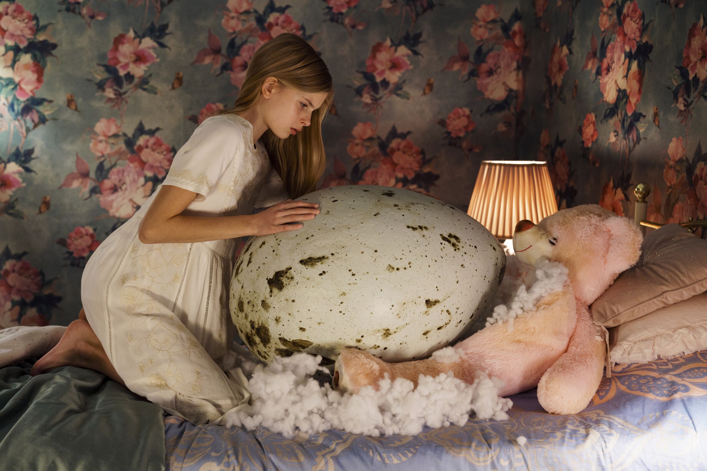 A girl looks over a giant egg in a bed with a large teddy bear 