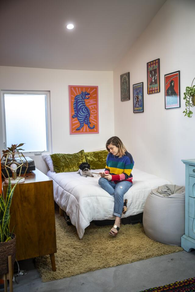 A woman and dog sit on a bed, with a dresser alongside.