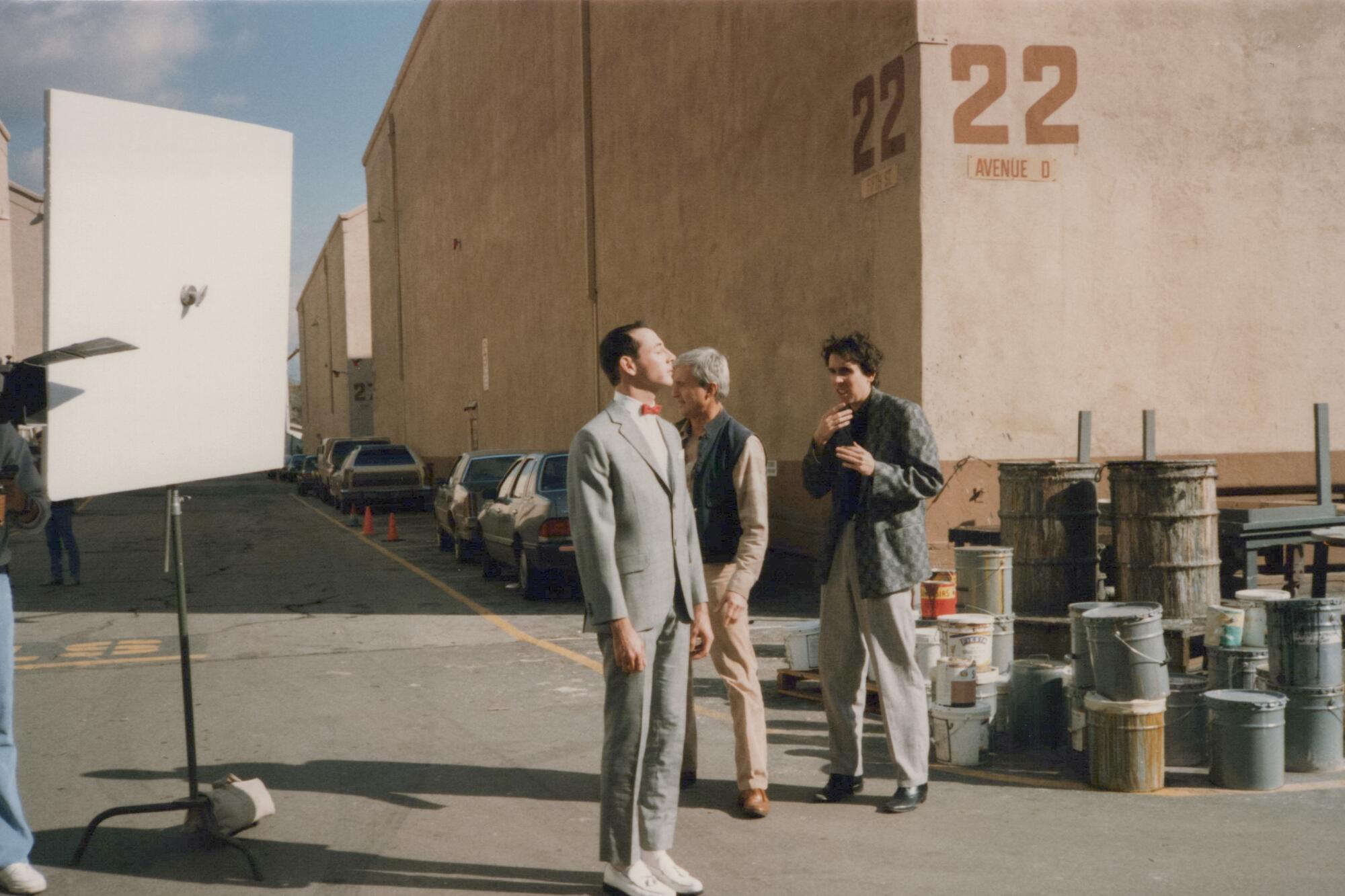 A man in gray suit, red bowtie and white shoes stands at intersection of a studio lot alleyway; two people walk behind him.