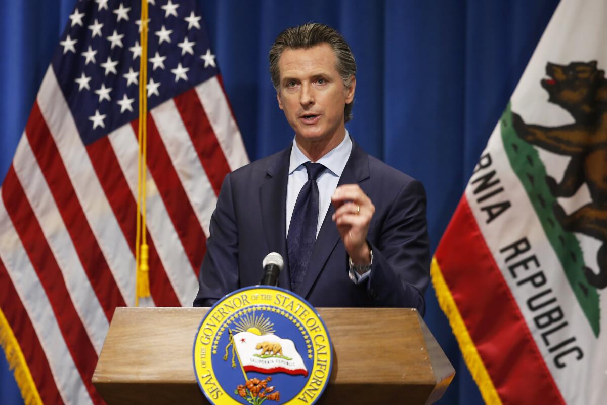 Gavin Newsom gestures with his left hand while speaking at a lectern in front of U.S. and California flags