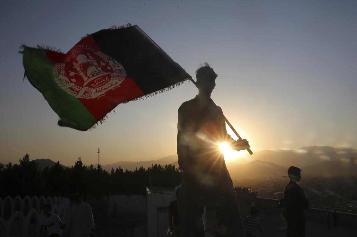 A silhouetted figure backlit by the setting sun waves a tattered Afghan flag.