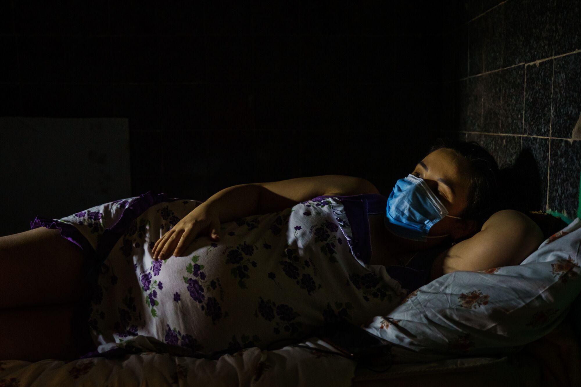 A pregnant woman with a face mask on lying on her side in a darkened shelter
