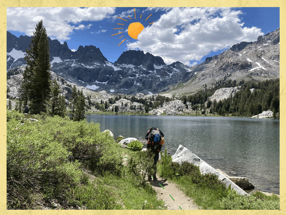 A hiker with a backpack walks on a trail next to a still lake backed by mountains.