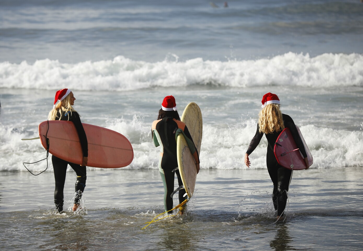 Wearing Santa hats, Crissy Seggerman, left, Heather Nagey, and Kaitlyn Johnson walk out to surf during the 9th Annual URT Santa Surf Off and Toy Drive in Coronado on Dec. 24, 2019.