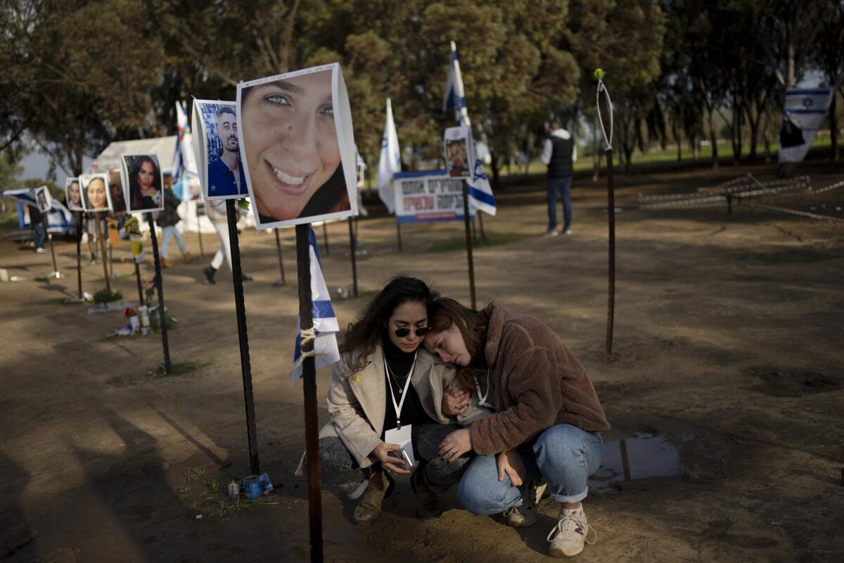 Two women sit next to a small pole holding an Israeli flag and a portrait of a young woman, among rows of such poles. 