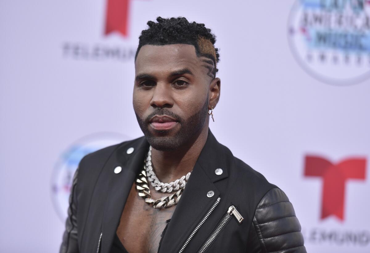 Jason Derulo in a black Moto jacket and large chains posing at a red carpet. 
