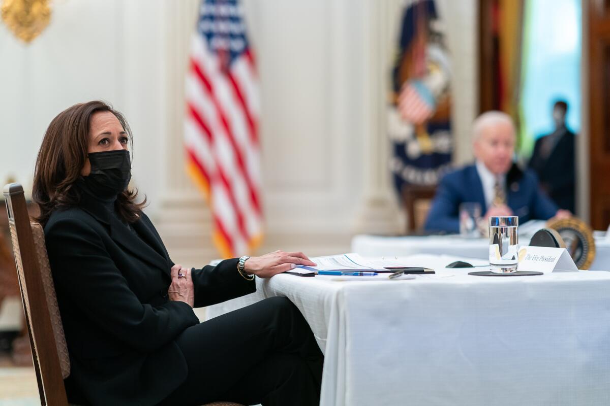 Kamala Harris wears a mask and sits at a table. Joe Biden sits at another table in the background.