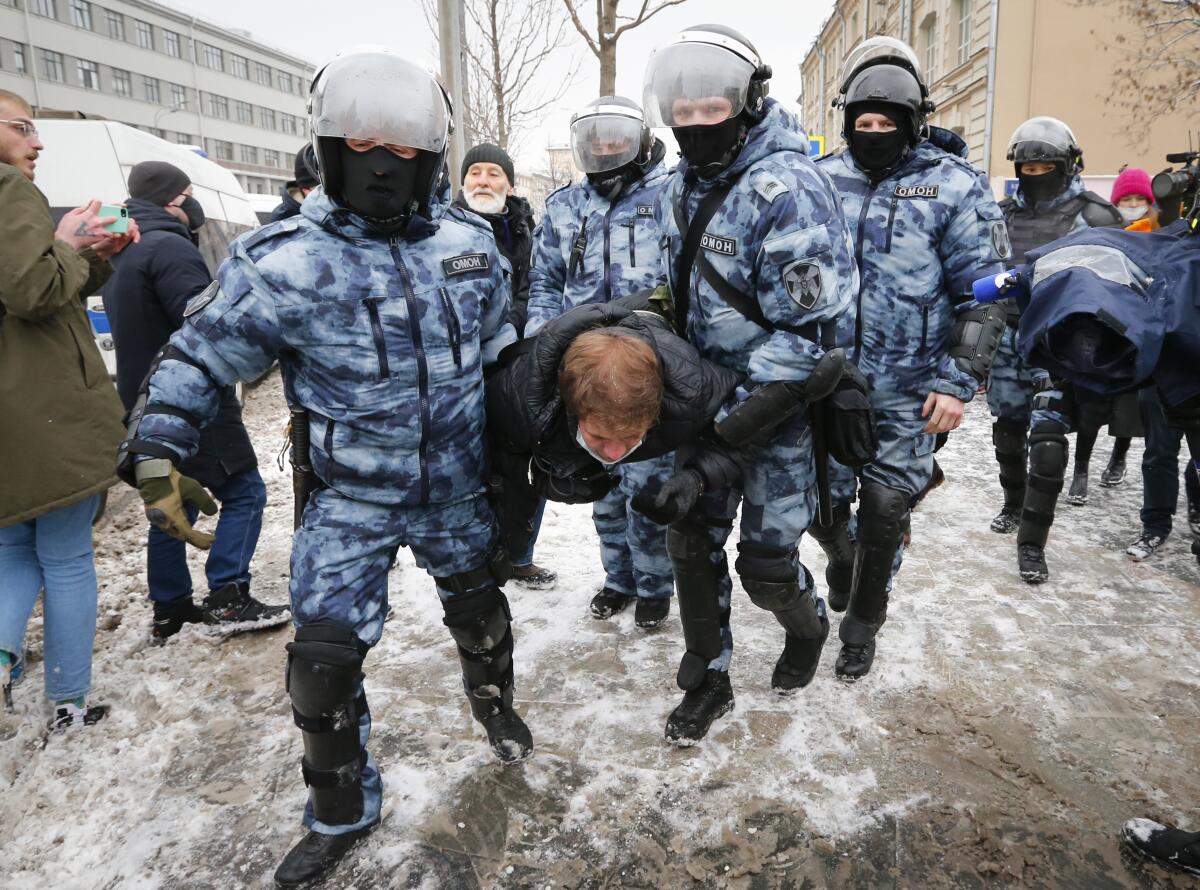 A man is carried away by several men in uniforms and helmets across dirty snow.