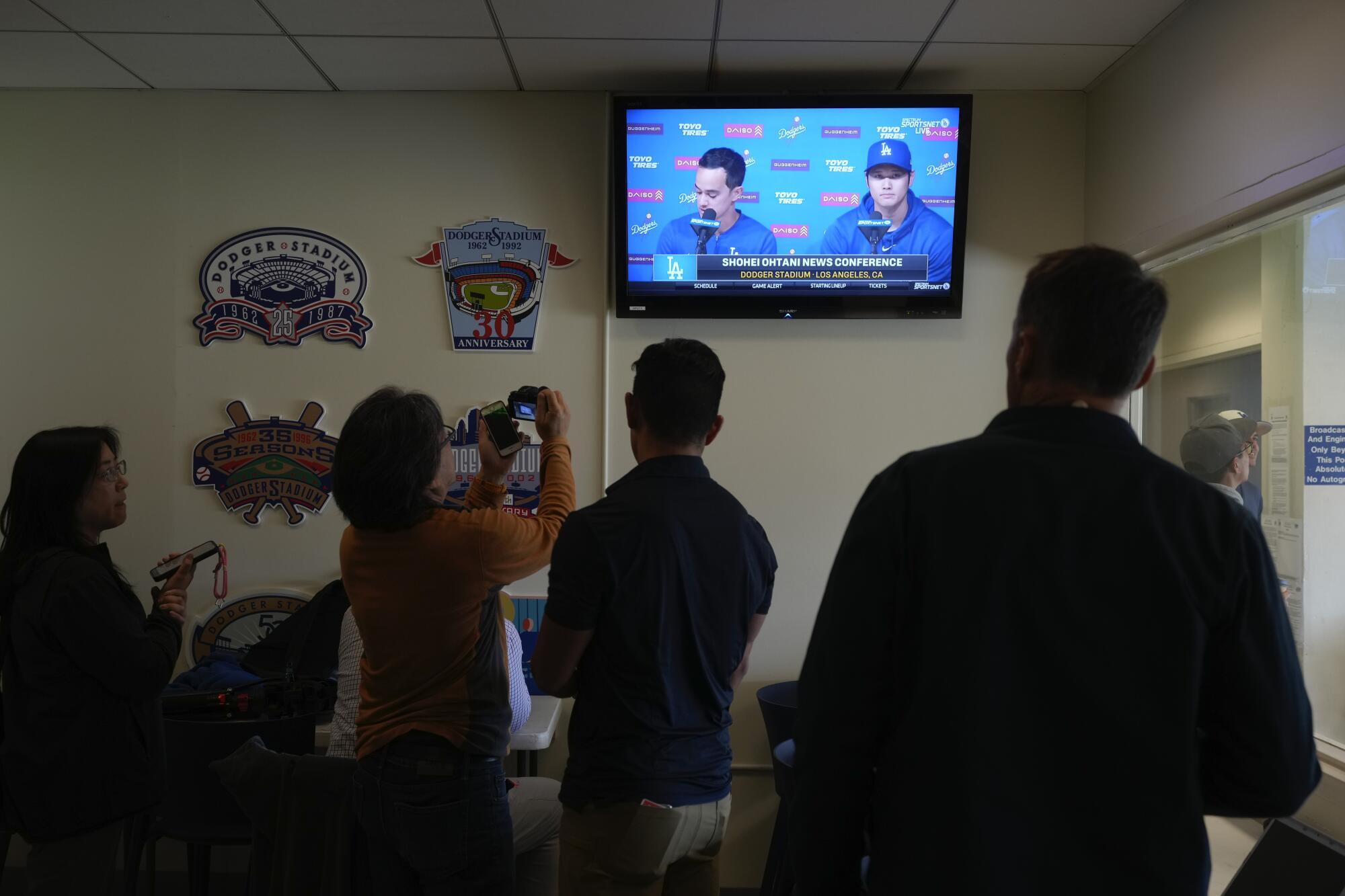 Reporters watch the TV showing Dodgers star Shohei Ohtani, right, and his new interpreter, Will Ireton, at a news conference.