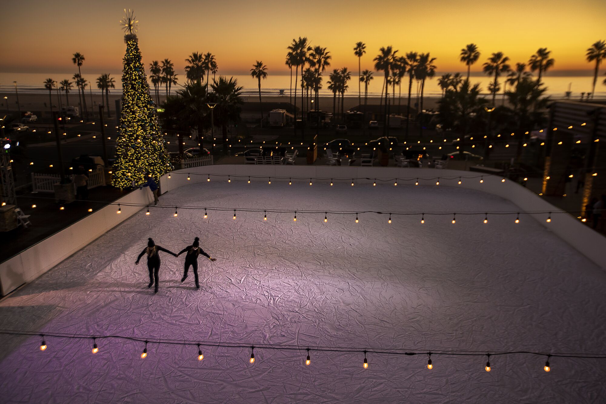 Two people ice skate on an outdoor rink. In the background is a lighted Christmas tree, palm trees and the ocean.