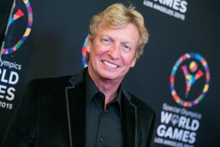 Nigel Lythgoe, with dirty blond hair, smiles while posing in a black suede suit jacket and black dress shirt 