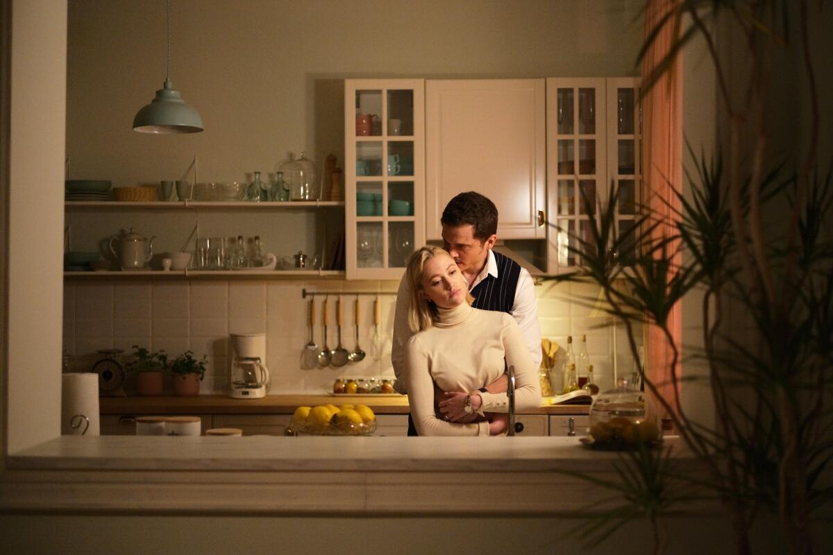 A man embraces a woman from behind in a kitchen in the movie “Watcher.”