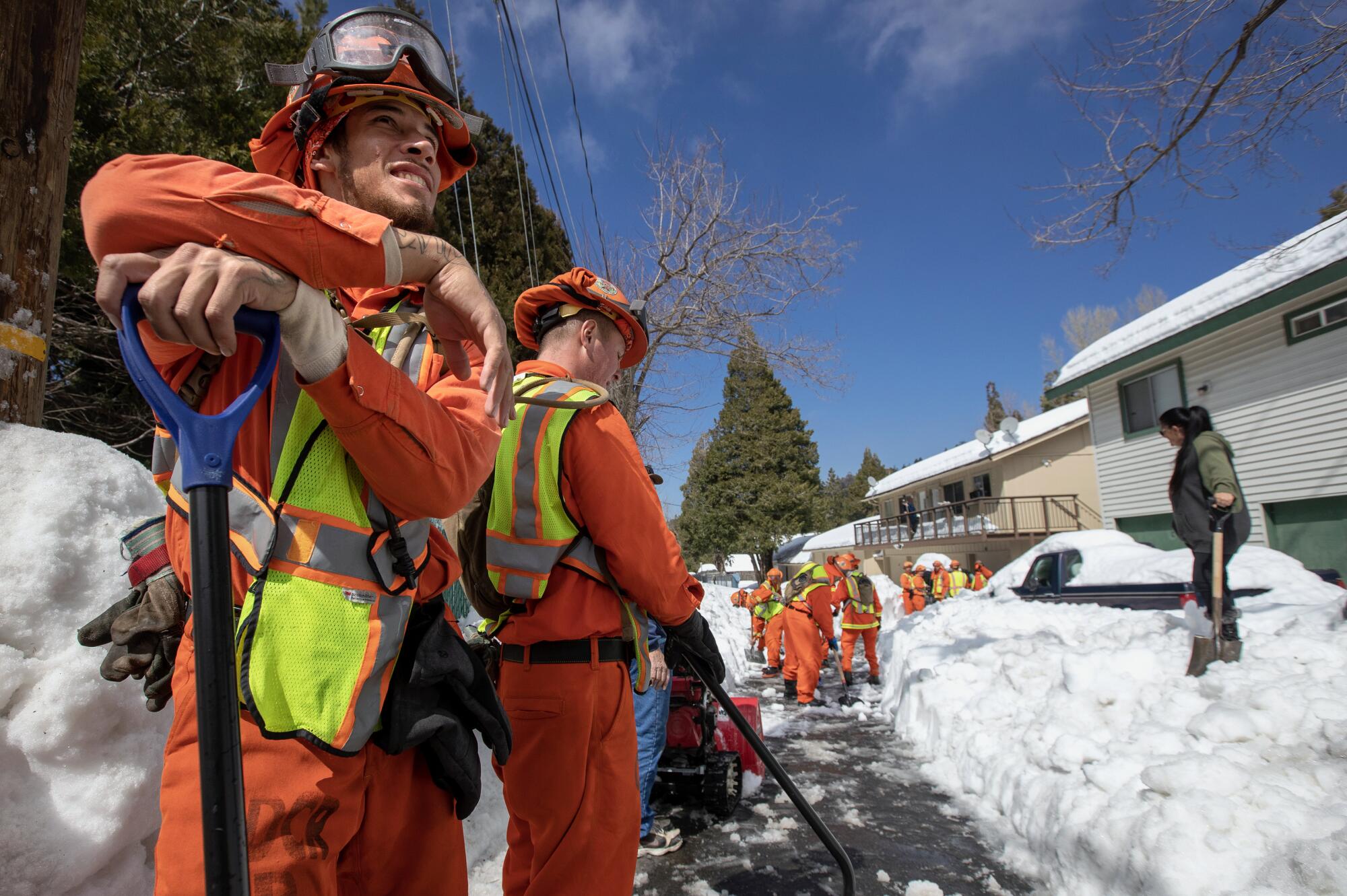 Men in orange jumpsuits, orange helmets and reflective vest dig with snowshovels along a pathway in front of two homes 