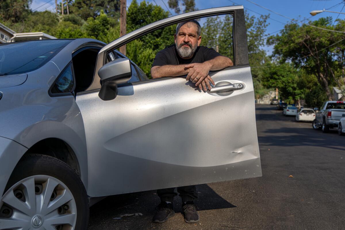 A man holds open a dented driver's side door of a car parked on a street.