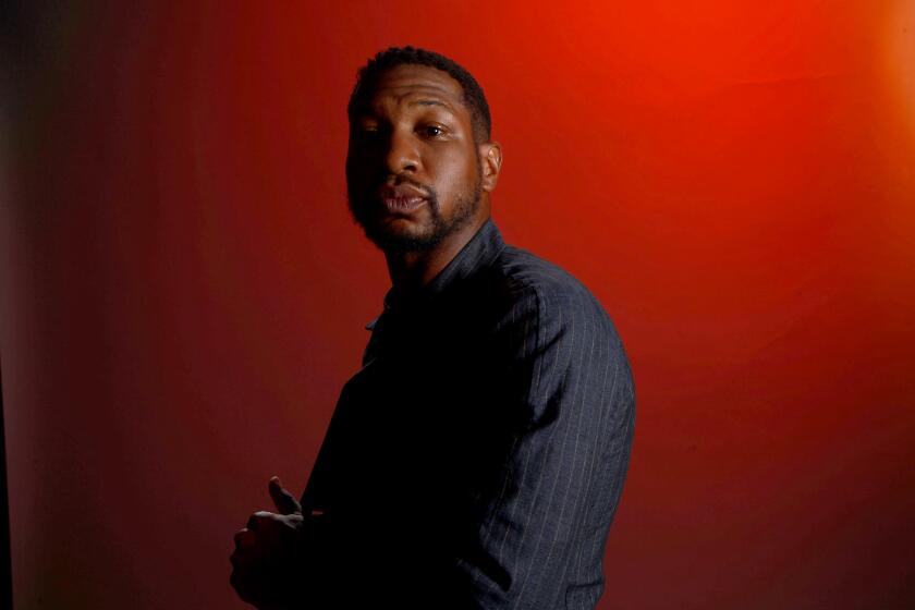Jonathan Majors in a long-sleeve shirt posing at a side-profile against a dark red background with shadows in a corner