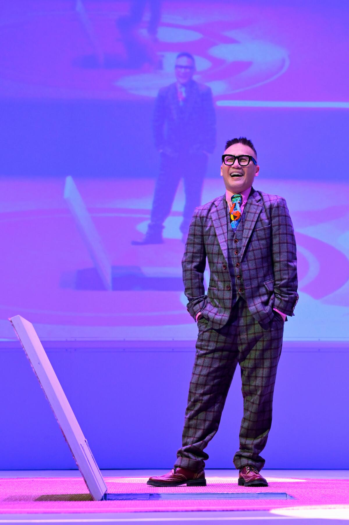 A smiling person in a checked suit and multicolored tie stands on a stage with a screen behind him showing him.