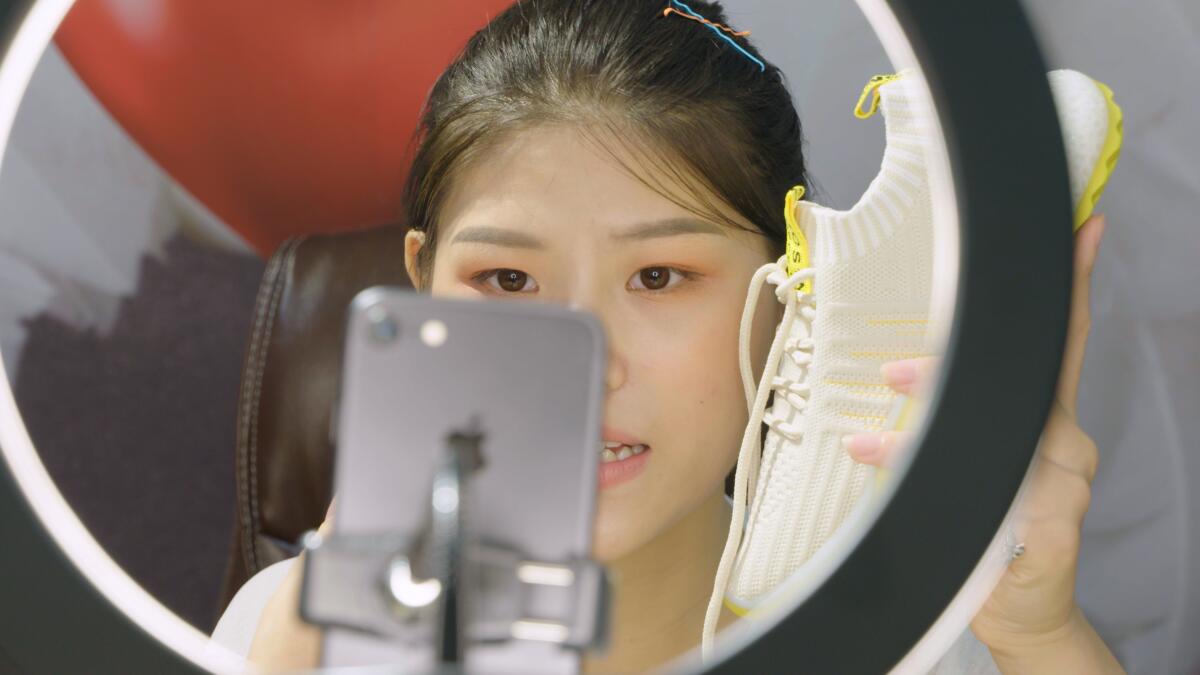 A woman holding a shoe sits in front of a cellphone and a ring light.