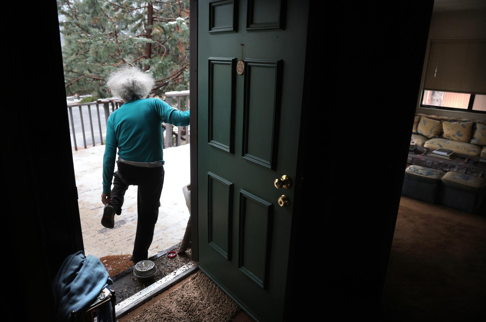 A man with gray hair, in a turquoise shirt and dark pants, heads out his front door into a snowy landscape with trees 