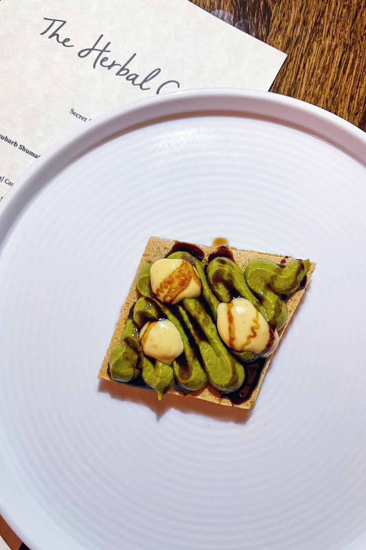 A round plate containing a square cracker topped with leeks and foie gras.