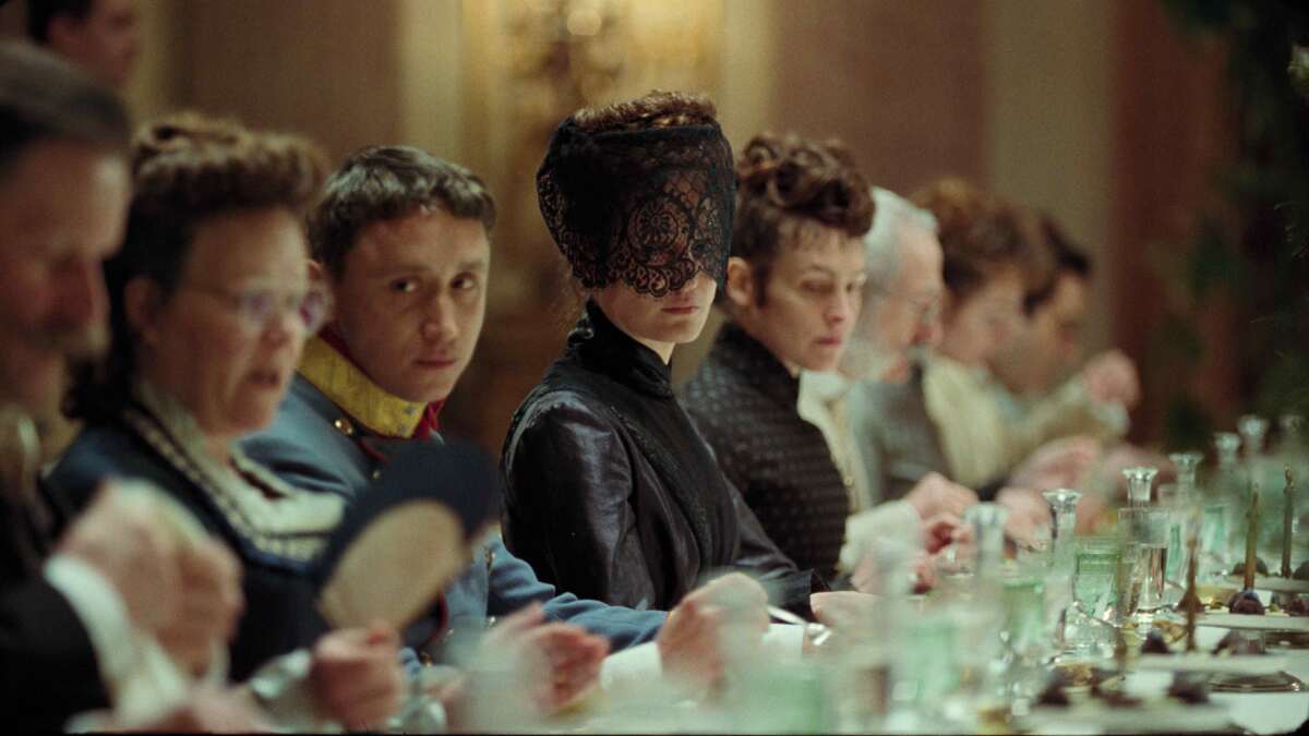 A masked woman and other people in Victorian evening dress sit at a table in the movie "Corsage."