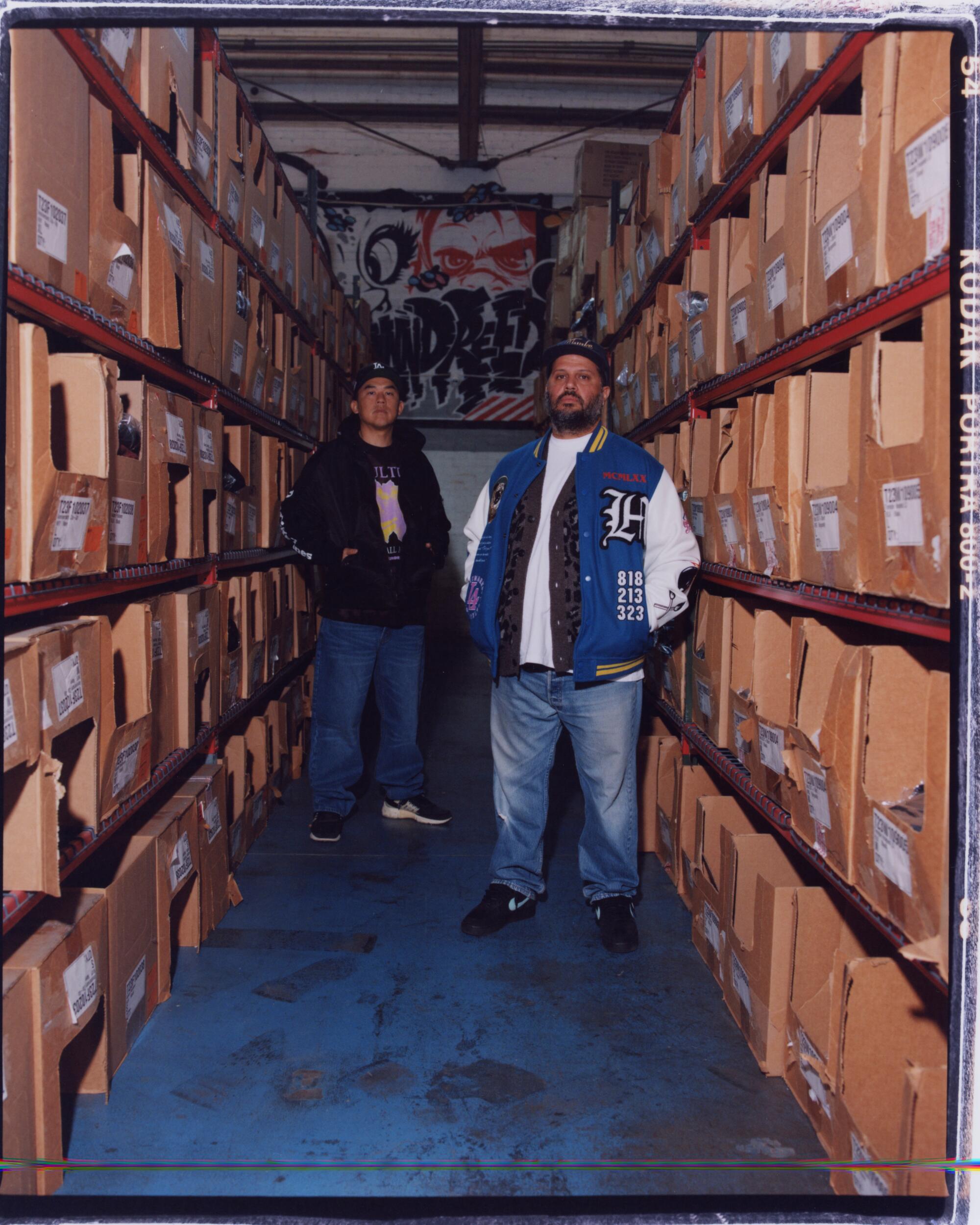 Two man stand between tall shelves filled with cardboard boxes