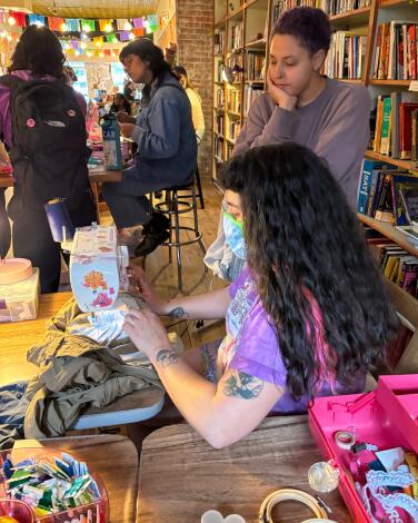 People at a crafts table with a sewing machine, one is sitting and the other is standing.