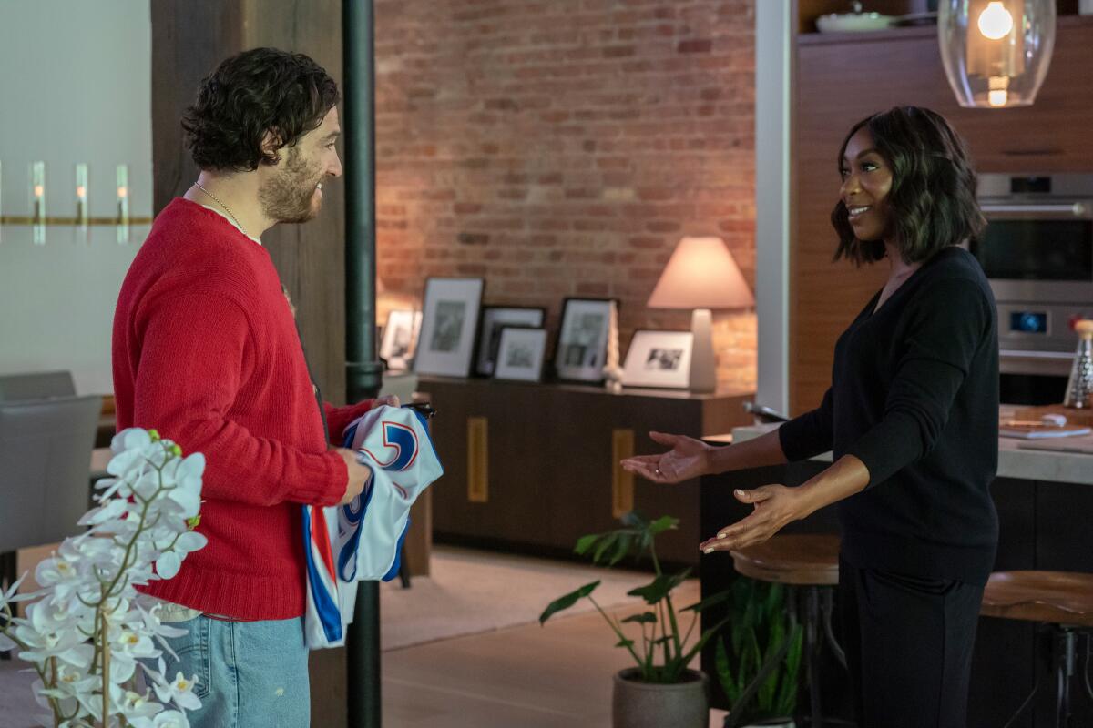 A man in a red sweater holding a jersey looks at a woman in dark clothing.