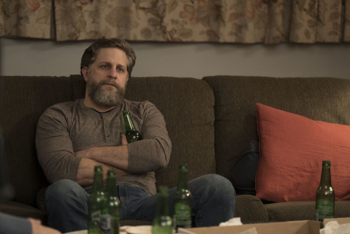 A man sits on the couch with his arms crossed, empty beer bottles littered around him