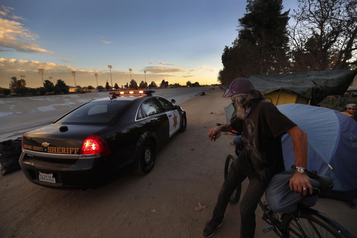 A man sitting on a bike net to a homeless encampment along a river bed watches a sheriff's cruiser drive past
