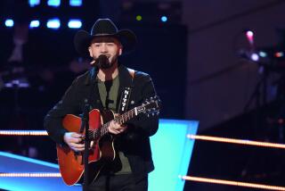 A. man in a Cowboy hat, a blazer and a grey shirt holding a guitar and singing on a stage