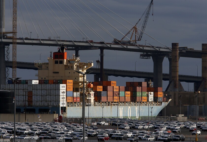 A loaded cargo container ship looms beside cars in a parking lot.