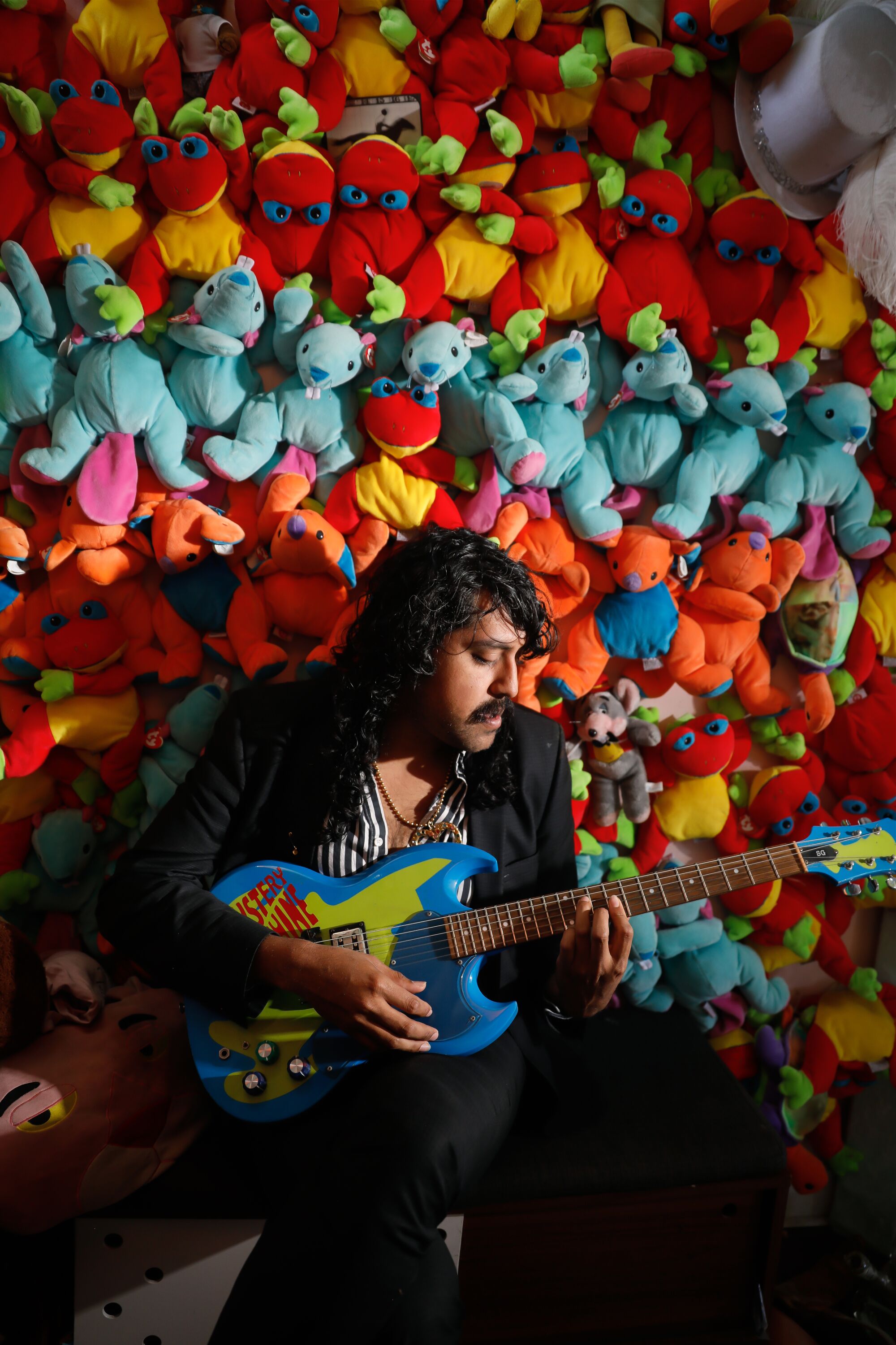 Enrique Tena Padilla (aka DJ Escuby), in a black suit, strums a guitar before a wall covered in Beanie Babies.