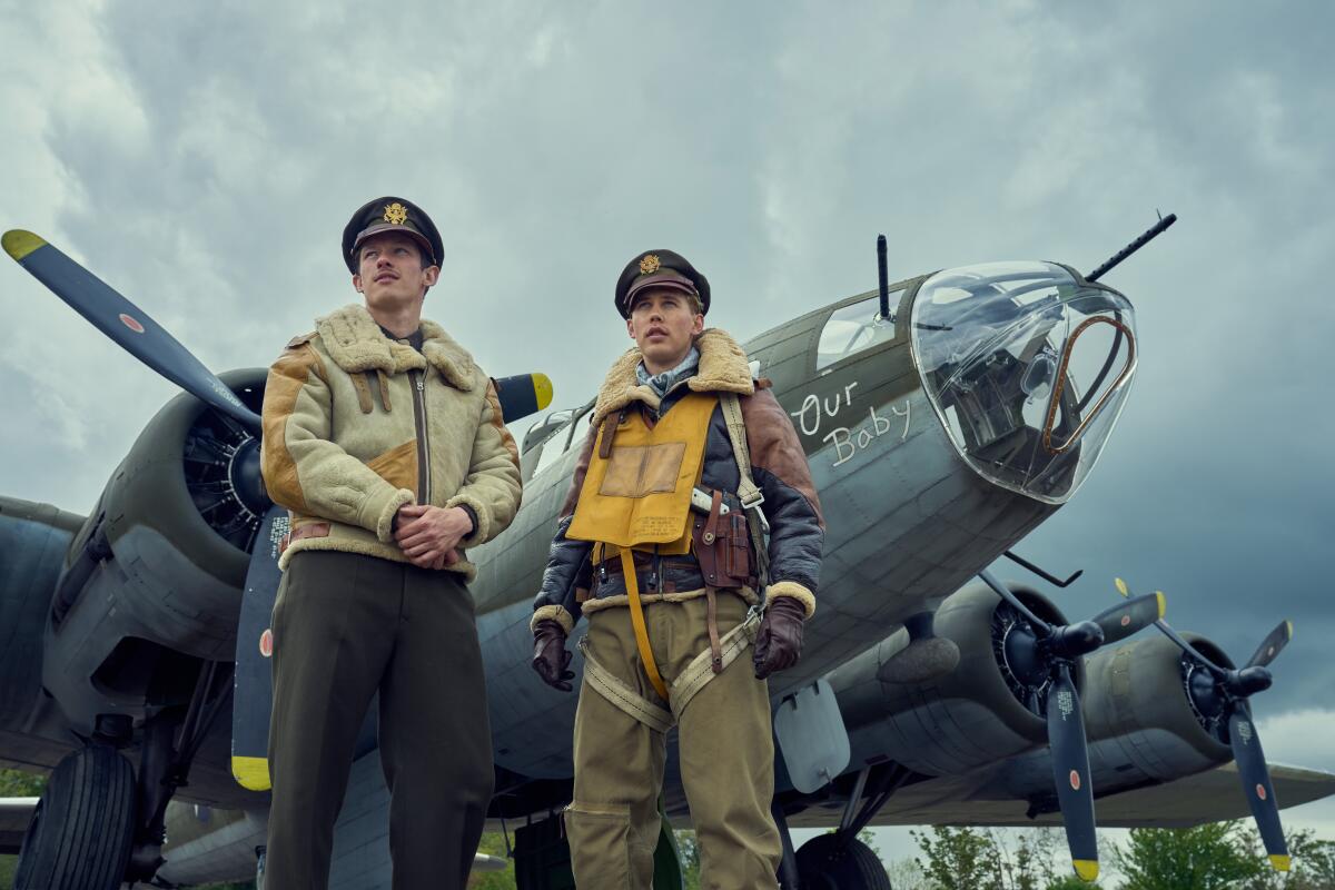 Two men in WWII military uniforms stand in front of a propeller plane of the era in "Masters of the Air." 