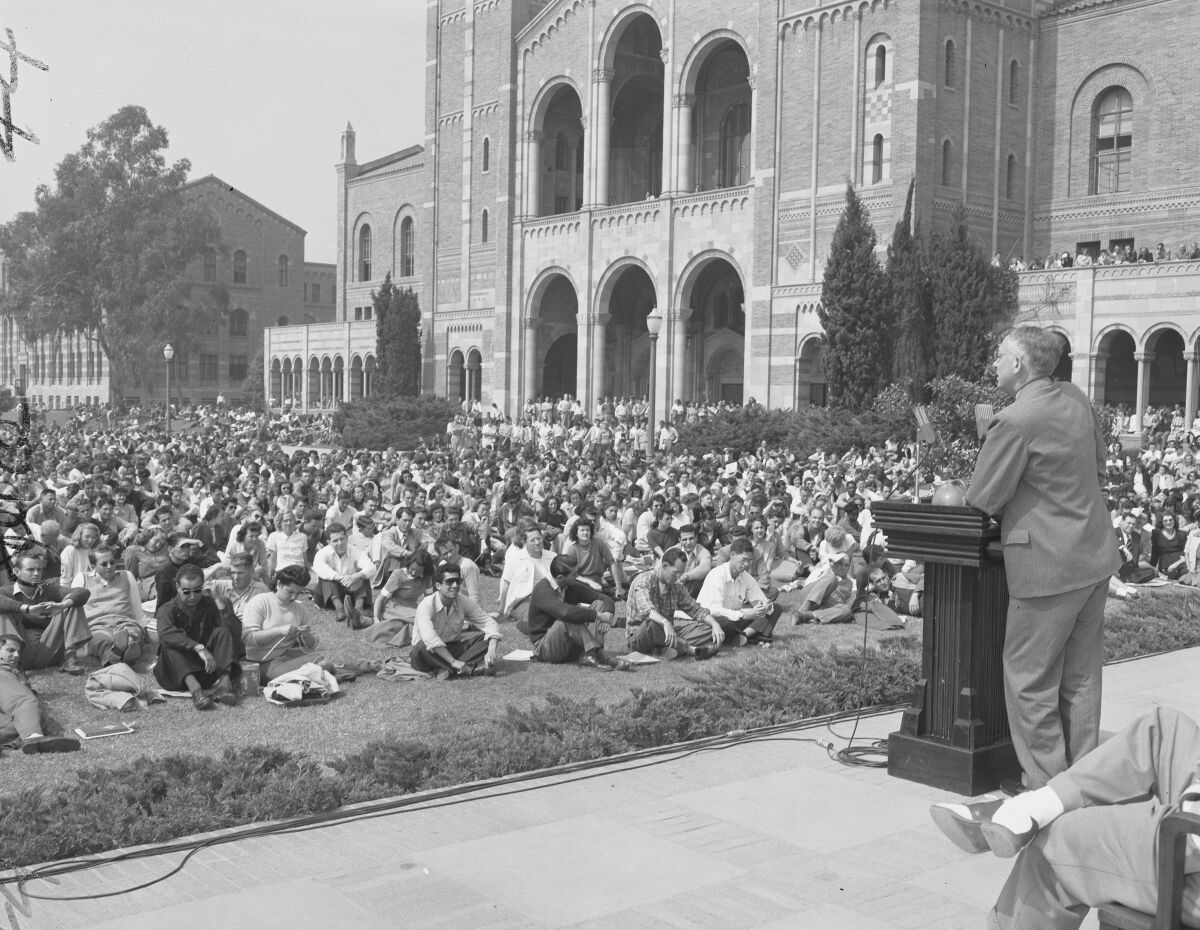 A man in a suit speaks at a lectern. Young people sit in rows on the grass in front of an imposing building.