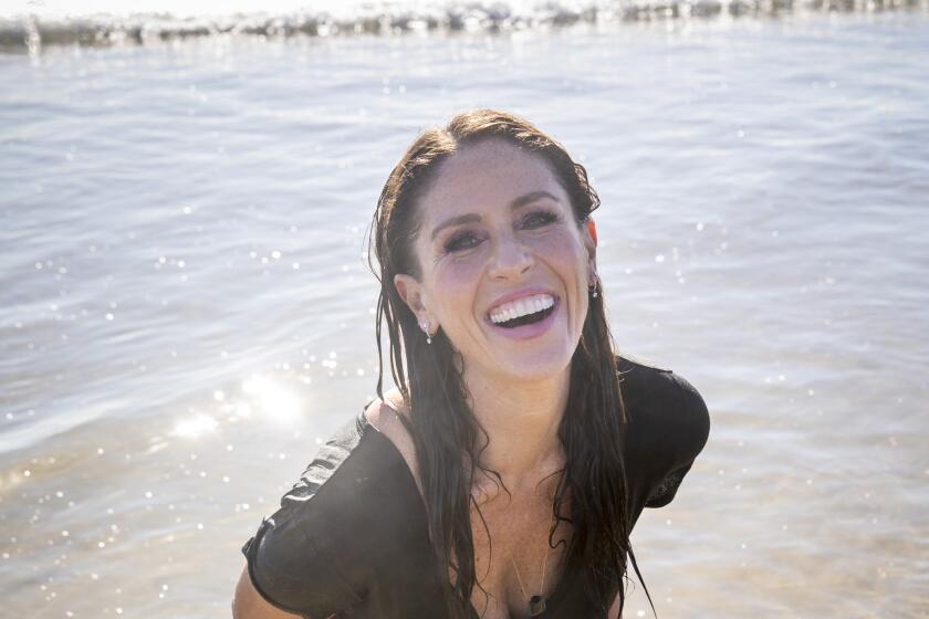 A smiling woman standing in the water in a brown t-shirt and blue jeans at the beach