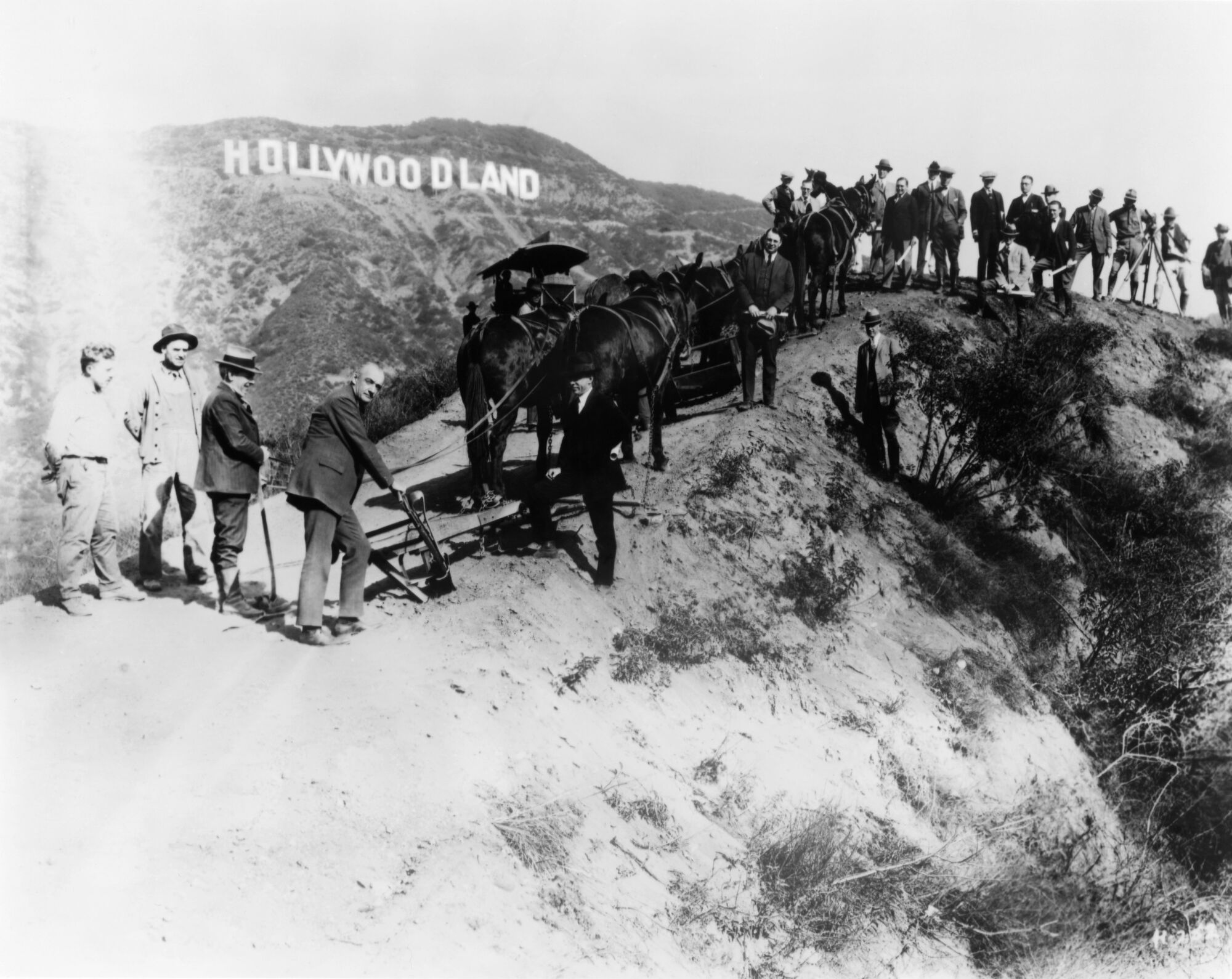 A group of men and pack animals on a hill with the "Hollywoodland" sign in the distance