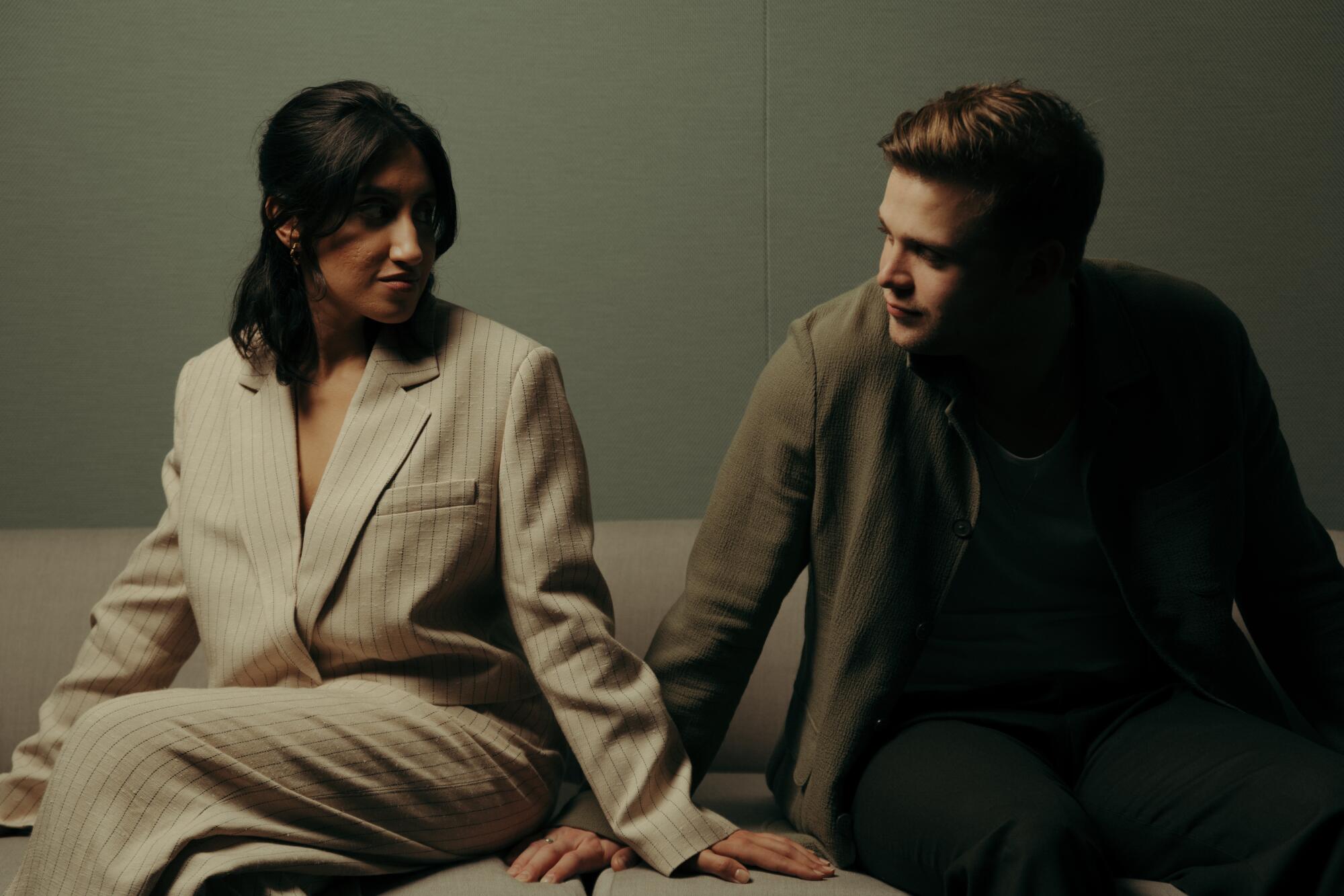 A woman in beige suit sits next to and looks at a man in a brown coat.