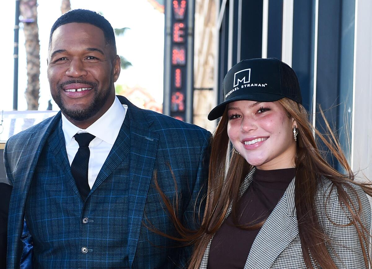 Michael Strahan in a blue plaid suit smiling next to a young woman with long hair and a black hat in a gray blazer