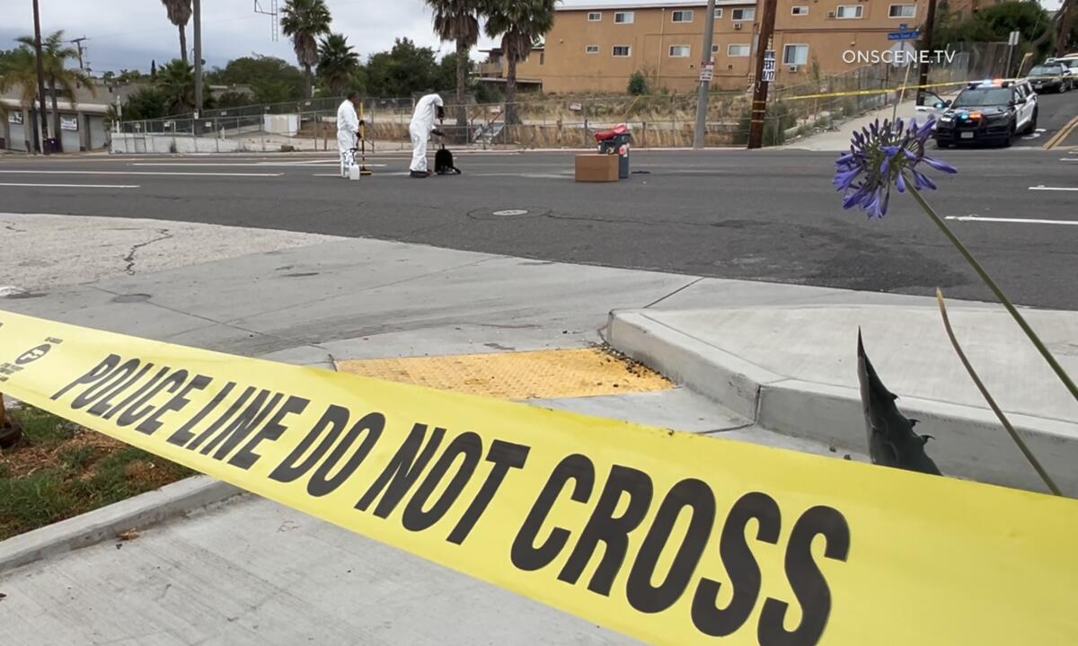 Yellow tape is seen in the foreground. In the background two men in white jumpsuits work in an empty roadway.
