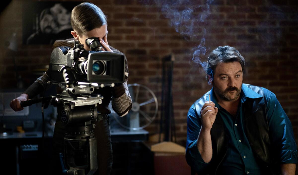 A man sits smoking next to a person working a movie camera.