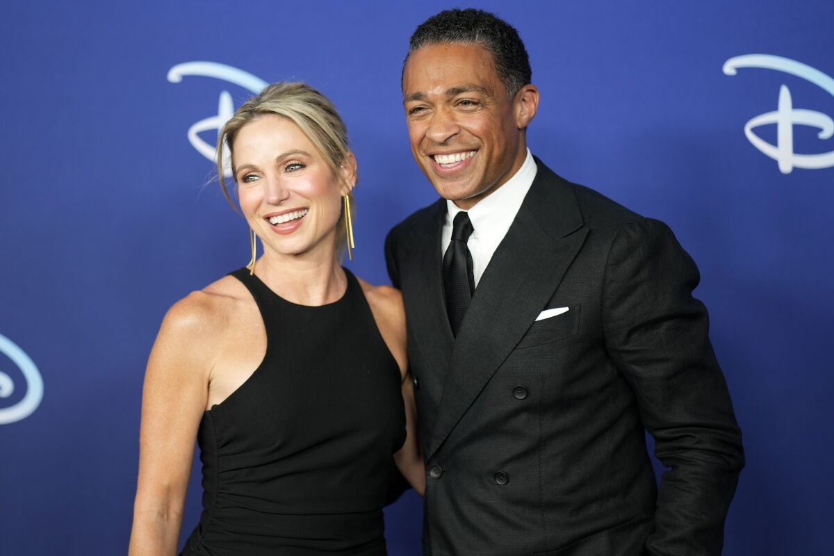 A blond woman in a black dress and a dark-haired man in a black suit smile and pose for a photo together