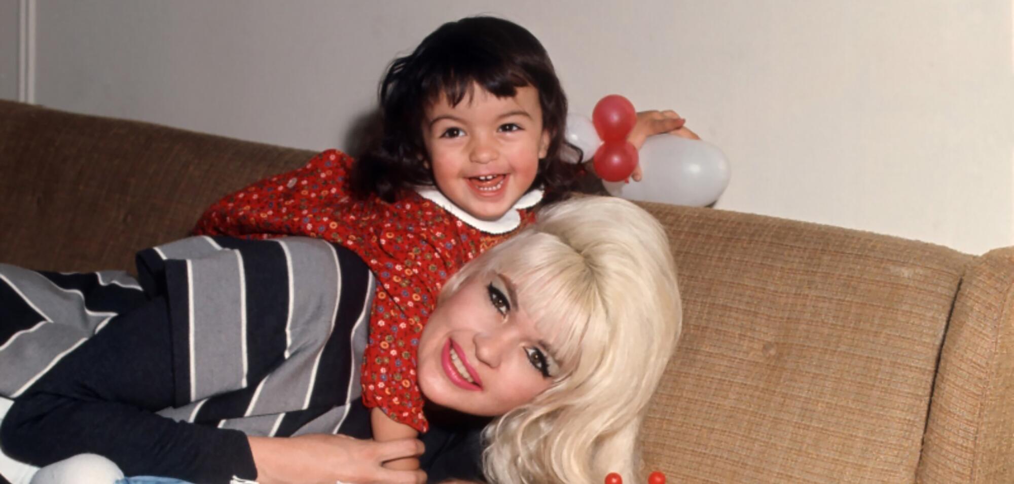 A little girl in a red dress leans on the back of a blond smiling woman.